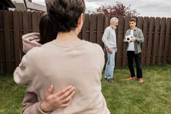 Middle aged man standing near young son with football near blurred wife and daughter during parents day celebration at backyard, quality time with parents concept, tradition and celebration — Stock Photo