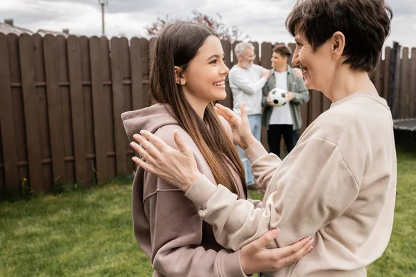 Cheerful middle aged mother hugging teenage daughter near blurred family with football standing at backyard during parents day celebration in june, quality time with parents concept — Stock Photo