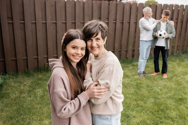 Smiling middle aged woman hugging teenage daughter and looking at camera near blurred family with football during parents day celebration at backyard, quality time with parents concept — Stock Photo