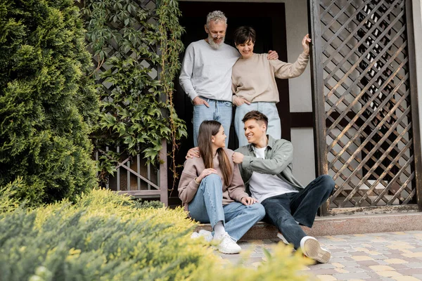 Cheerful middle aged parents hugging and looking at children sitting on porch of house during parents day celebration at backyard, family traditions and celebrations concept — Stock Photo