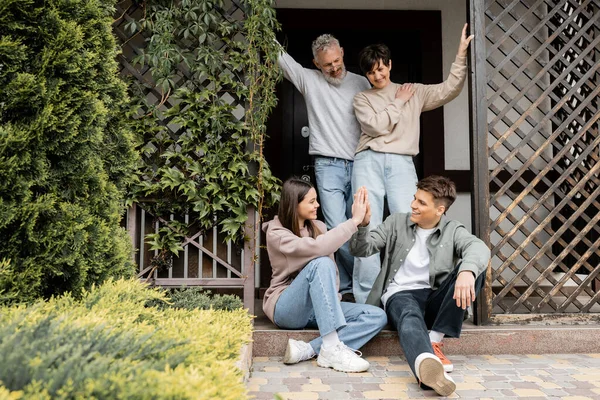 Positive middle aged parents looking at children giving high five and standing on porch of house during parents day celebration in june, family traditions and celebrations concept, special occasion — Stock Photo