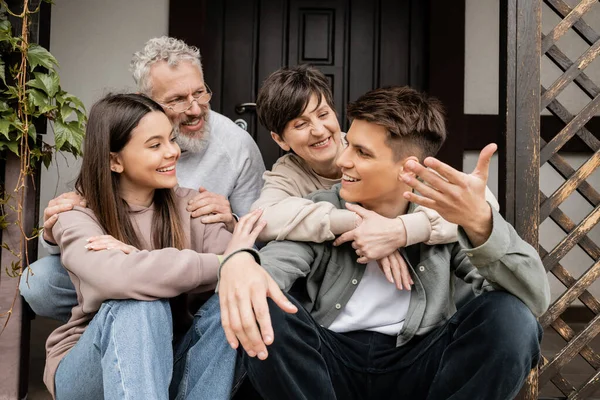Усміхнений молодий чоловік розмовляє з сім'єю і сестрою, сидячи на ганку будинку під час святкування дня батьків у червні, сімейні традиції та концепція святкування, особливий випадок — стокове фото