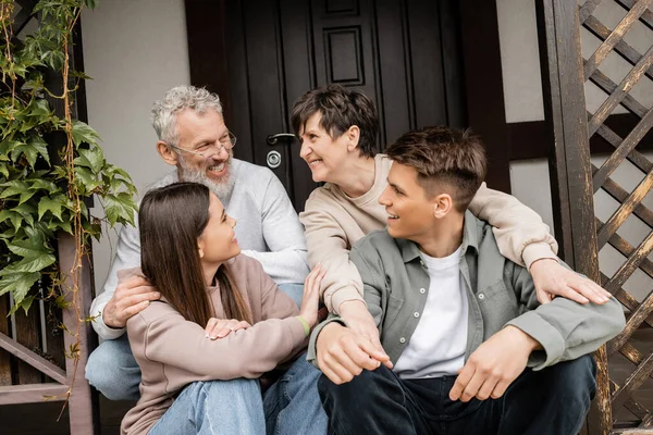 Parents d'âge moyen positifs parler tout en embrassant les enfants et assis sur le porche de la maison lors de la célébration de la journée des parents en juin, traditions familiales et concept de célébrations — Photo de stock