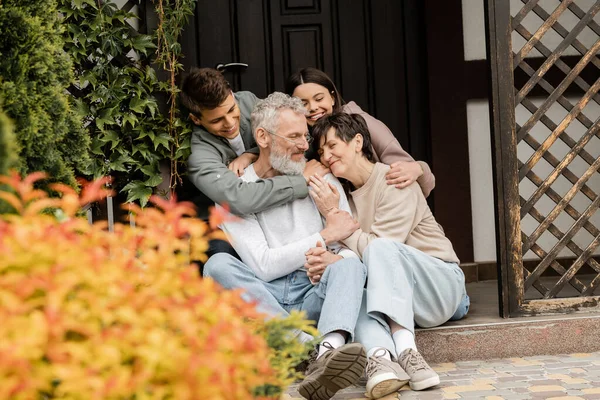 Lächelnde Kinder umarmen glückliche Eltern mittleren Alters, während sie gemeinsam auf der Veranda des Hauses sitzen und im Juni den Elterntag im Hinterhof feiern, Familientradition und Festkonzept — Stockfoto