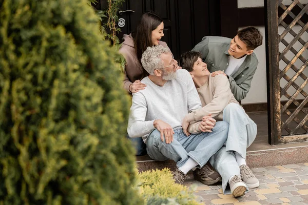 Cheerful kids hugging and talking to middle aged parents while sitting together on porch of house and celebrating parents day in june, family traditions and celebrations concept — Stock Photo