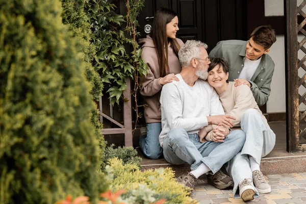 Smiling middle aged parents holding hands while sitting on porch of house near kids and celebrating parents day at backyard, family traditions and celebrations concept, special occasion — Stock Photo