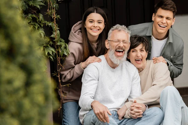 Positive children looking at camera while hugging middle aged parents holding hands and sitting on porch of house during parents day celebrating , parent-child relationship concept — Stock Photo