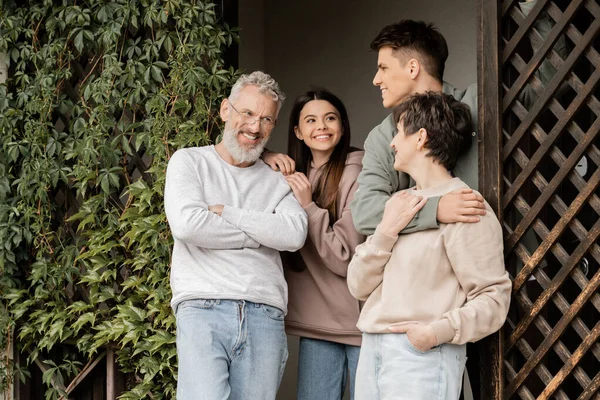Cheerful young man hugging middle aged mother near father and teenage sister while standing on porch of house and celebrating parents day in june, parent-child relationship concept — Stock Photo