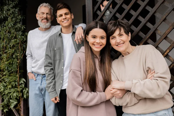 Smiling teenage girl holding hand of middle aged mother while looking at camera near blurred father and brother during parents day celebration on porch of house, parent-child relationship concept — Stock Photo