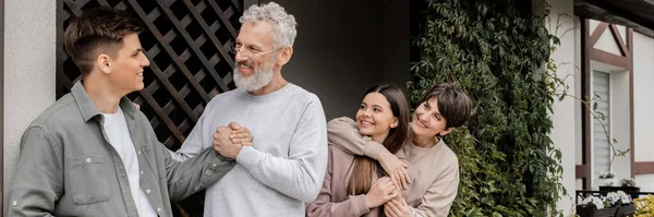 Homme d'âge moyen souriant serrant la main du jeune fils tandis que la femme étreignant sa fille lors de la célébration de la journée des parents près de la maison dans la cour, concept de relation parent-enfant, bannière — Photo de stock