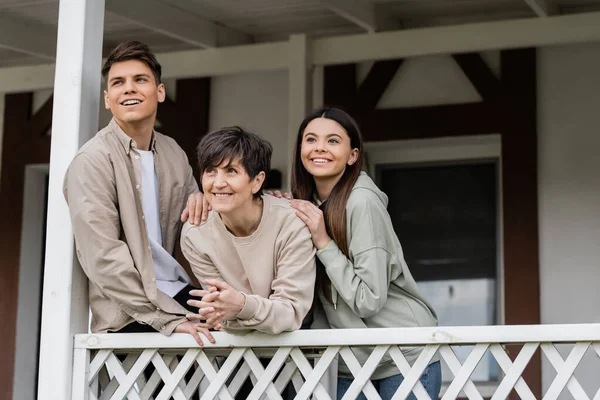 Positive siblings looking away while hugging middle aged mother and celebrating parents day, standing on porch of house in june, parent-child relationship concept, special occasion — Stock Photo