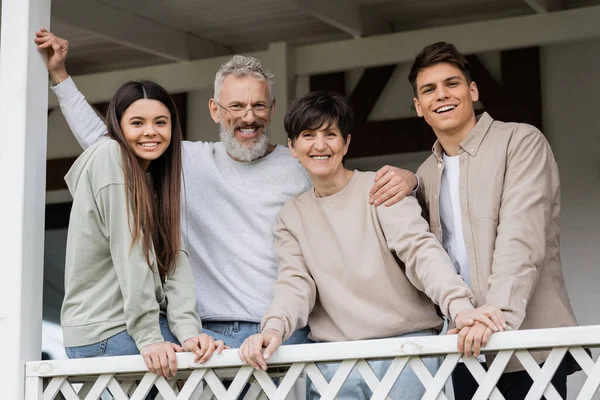 Portrait of positive middle aged parents looking at camera while standing near kids and celebrating parents day near house on porch, parent-child relationship concept, special occasion — Stock Photo