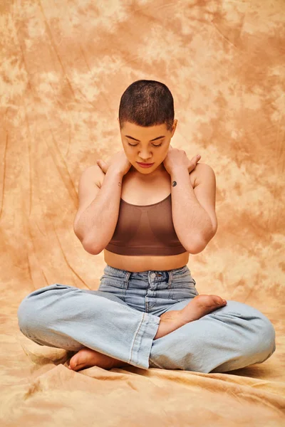 Body confidence, relaxation, curvy young and tattooed woman in jeans and crop top sitting with crossed legs on mottled beige background, personal style, self-acceptance, generation z, denim fashion — Stock Photo