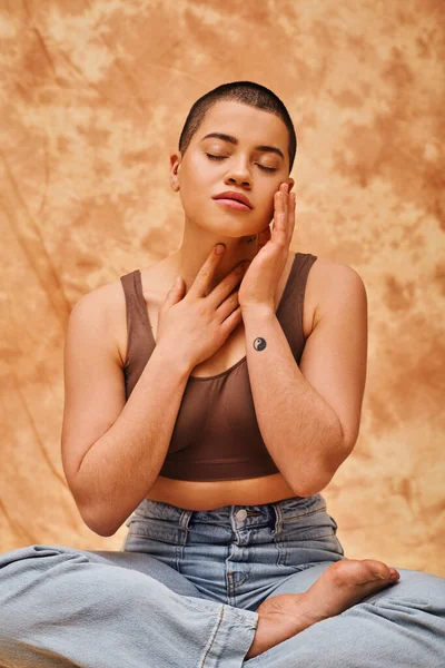 Body image, relaxation, curvy young and tattooed woman in jeans and crop top sitting with crossed legs on mottled beige background, closed eyes, personal style, self-acceptance, generation z — Stock Photo