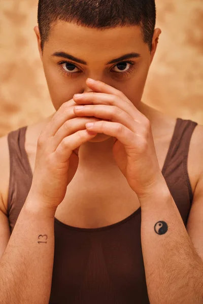 Positividad corporal, mujer curvilínea y tatuada mirando a la cámara sobre fondo beige moteado, manos cerca de la cara, autoaceptación, generación z, cultura juvenil, pelo corto y joven, retrato - foto de stock