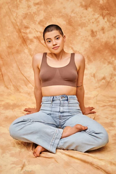 Body positivity, casual attire, curvy young and tattooed woman in jeans and crop top sitting with crossed legs on mottled beige background, personal style, self-acceptance, generation z — Stock Photo