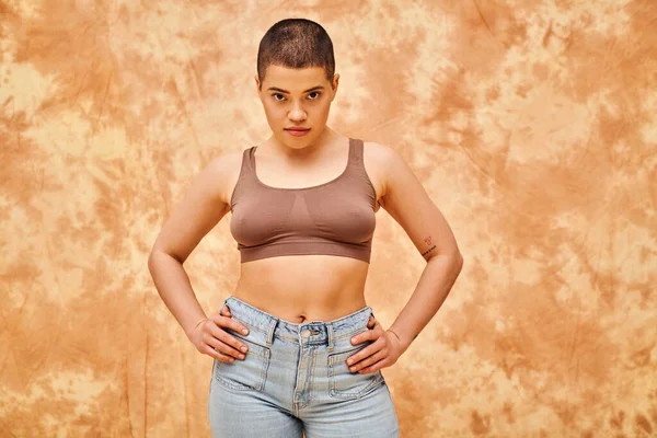 Body positivity movement, curvy young woman with tattoos posing in jeans and crop top on mottled beige background, hands on hips, representation of body, confidence, casual attire, generation z — Stock Photo