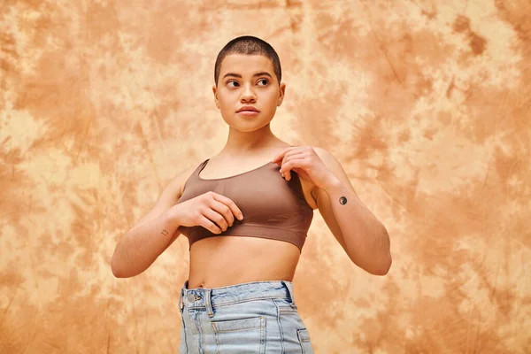 Body positivity movement, curvy young woman with tattoos posing in jeans and crop top on mottled beige background, representation of body, confidence, casual attire, generation z, looking away — Stock Photo