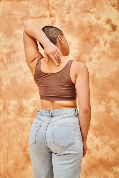 Curvy model, back view of young short haired woman standing with hand behind back, mottled beige background, representation of body, different shapes, generation z, youth, body positivity — Stock Photo