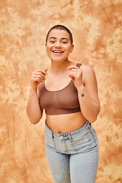 Body positivity, happy and tattooed woman in crop top and jeans posing on mottled beige background, looking at camera, representation of body, different body shapes, generation z, youth, tattooed — Stock Photo