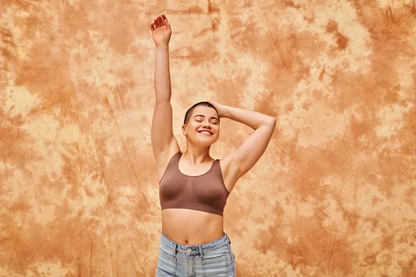 Body positivity, curvy and cheerful woman in crop top posing on mottled beige background, smiling with closed eyes, raised hand, representation of body, body shapes, generation z, youth, tattooed — Stock Photo