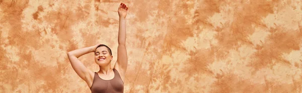 Body positivity, curvy and tattooed woman in crop top posing on mottled beige background, smiling with closed eyes, raised hand, representation of body, body shapes, generation z, youth, banner — Stock Photo