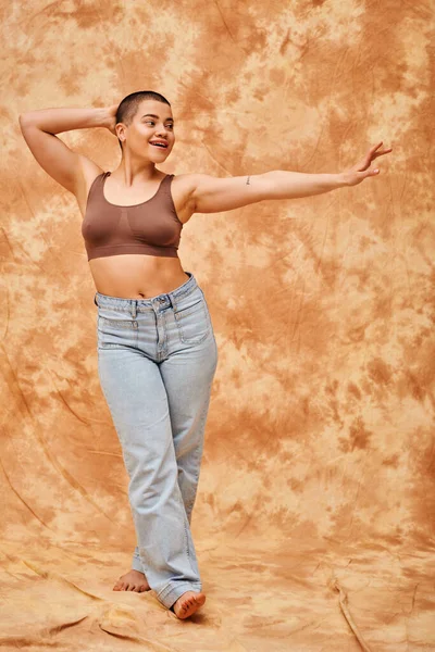 Body positivity, jeans look, curvy and happy woman in crop top posing with outstretched hand on mottled beige background, casual attire, looking away, self-acceptance, generation z, tattooed — Stock Photo