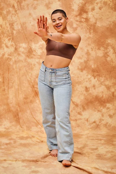 Движение тела позитивность, джинсы взгляд, кривая и радостная женщина в кукурузном топе позирует с протянутыми руками на пятнистом бежевом фоне, случайный наряд, принятие себя, поколение z, татуировка — стоковое фото