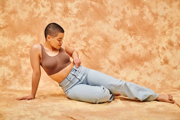 Позитивность тела, уверенная, пышная и молодая женщина в кукурузном топе и джинсах, позирующая на бежевом фоне с пятнами, повседневная одежда, самопризнание, поколение z, татуировка, полная длина, джинсовая мода — стоковое фото