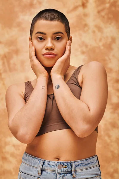 Body positivity and confidence, curvy young woman in crop top posing on mottled beige background, short haired, self-acceptance, generation z, tattooed, different shapes, looking at camera — Stock Photo