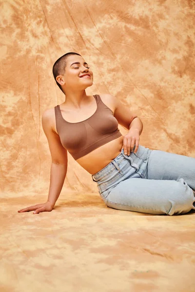 Body positivity, denim fashion, curvy and pleased woman in jeans and crop top sitting on mottled beige background, tattooed, casual attire, self-acceptance, generation z, body diversity — Stock Photo