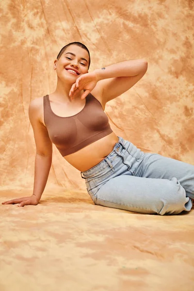 Body love, jeans look, curvy and tattooed woman in casual attire sitting on mottled beige background, confidence, self-acceptance, generation z, body diversity, pretty and positive - foto de stock