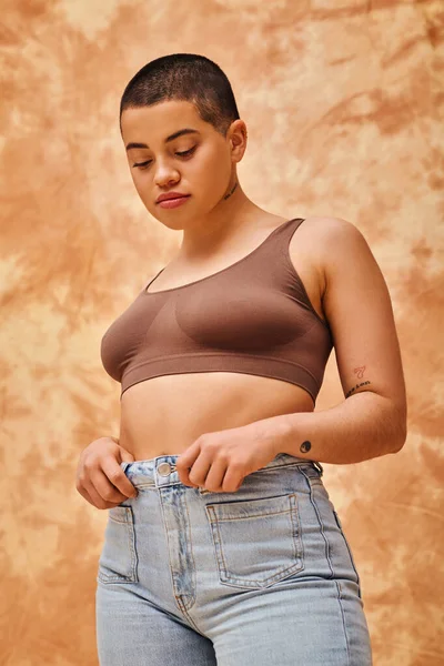 Body image, jeans look, curvy and tattooed woman in casual attire standing on mottled beige background, confidence, self-acceptance, generation z, body diversity, pretty and short haired model — Stock Photo