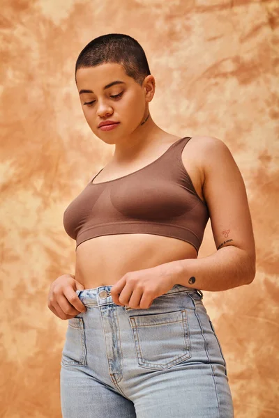 Image du corps, look jeans, femme courbée et tatouée en tenue décontractée debout sur fond beige marbré, confiance en soi, acceptation de soi, génération z, diversité du corps, modèle joli et à poils courts — Photo de stock