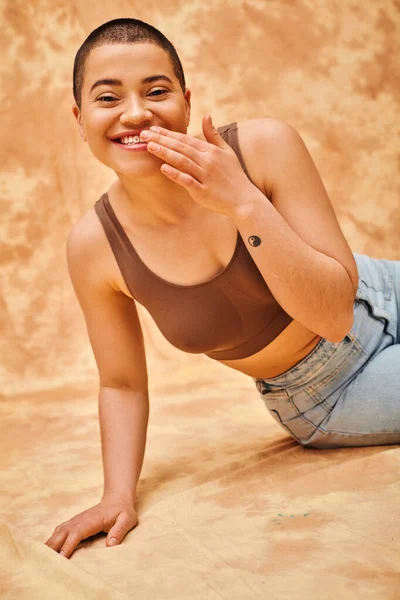 Denim fashion, gen z, joyful curvy woman with tattoo sitting on mottled beige background, body positivity movement, self-esteem, confidence, short haired model, youth culture, happiness — Stock Photo