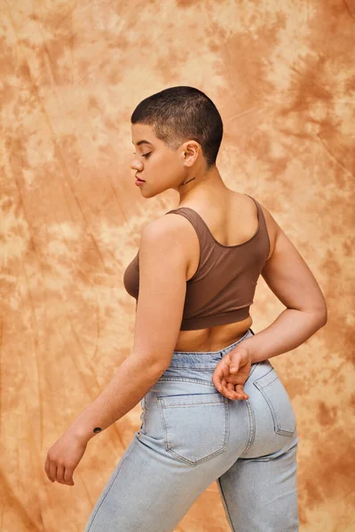 Denim fashion, gen z, curvy model with tattoo posing on mottled beige background, different shapes, body positivity movement, self-esteem, confidence, short haired woman, youth culture — Stock Photo
