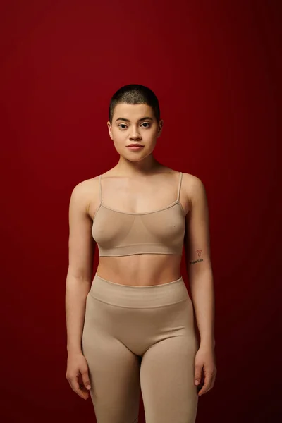 Body diversity, young tattooed woman in beige underwear posing on red background, body positivity, real people, burgundy, comfortable in skin, curvy model, generation z, self love, looking at camera — Stock Photo