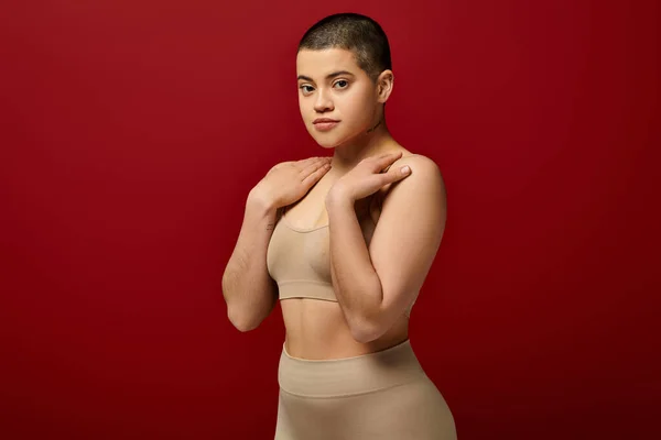 Body appearance, young tattooed woman in beige underwear posing on red background, body positivity, natural curves, comfortable in skin, curvy model, generation z, self love, looking at camera — Stock Photo