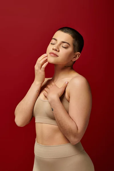 Body image, young tattooed woman in beige underwear posing on red background, body positivity, real people, burgundy, comfortable in skin, curvy model, generation z, self love, closed eyes — Stock Photo