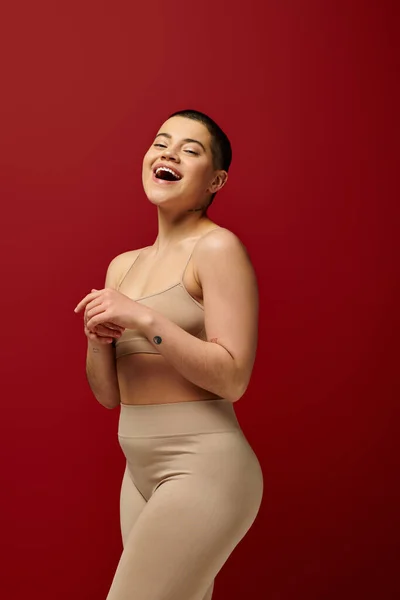 Self acceptance, amazed and tattooed woman in beige underwear posing on red background, body positivity, curvy fashion, comfortable in skin, curvy model, generation z, self love, short hair — Stock Photo