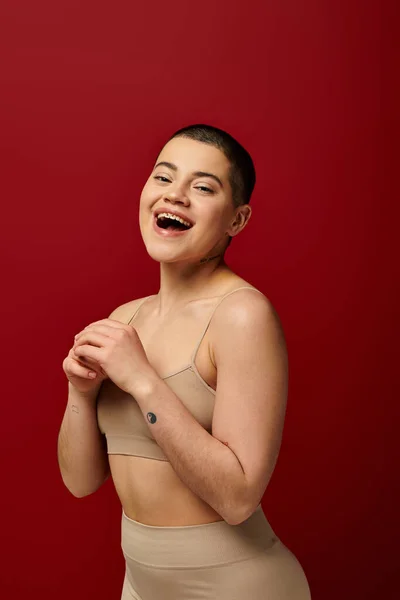 Self acceptance, amazed and tattooed woman in beige underwear posing on red background, body positivity, curvy fashion, comfortable in skin, self-acceptance, generation z, body diversity, laughter — Stock Photo