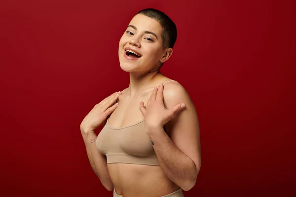 Self acceptance, excited and tattooed woman in beige underwear posing on red background, body positivity, curvy fashion, comfortable in skin, self-acceptance, generation z, body diversity, laughter — Stock Photo