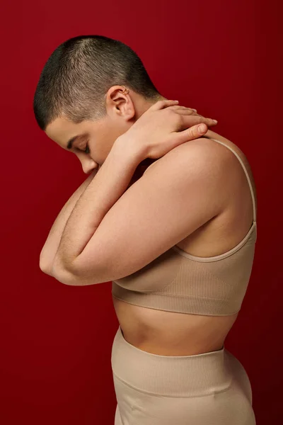 Selbstakzeptanz, Seitenansicht einer jungen Frau in beiger Unterwäsche, die auf rotem Hintergrund posiert, kurvige Mode, angenehm in der Haut, Selbstakzeptanz, Generation z, Körpervielfalt, kurze Haare — Stock Photo