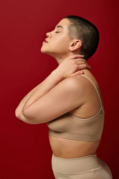 Self acceptance, side view of young woman with closed eyes posing  in beige underwear on red background, curvy fashion, comfortable in skin, self-acceptance, generation z, body diversity, short hair — Stock Photo
