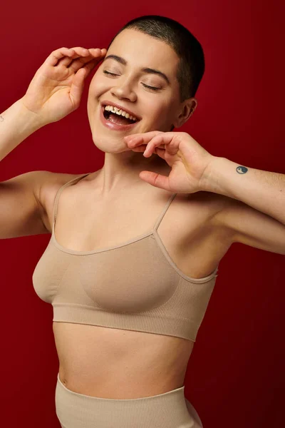 Self-esteem, excited and tattooed woman in beige underwear posing on red background, body positivity, curvy fashion, comfortable in skin, self-acceptance, generation z, body diversity, laughter — Stock Photo