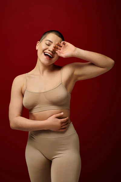 Selbstwertgefühl, glückliche und tätowierte Frau in beiger Unterwäsche, die auf rotem Hintergrund posiert, kurvige Mode, angenehm in der Haut, Körperpositivität, Selbstakzeptanz, Generation z, Körpervielfalt, Lachen — Stockfoto
