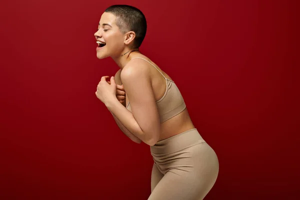 Selbstwertgefühl, glückliche und tätowierte Frau in beiger Unterwäsche, die auf rotem Hintergrund posiert, kurvige Mode, angenehm in der Haut, Körperpositivität, Generation z, Körpervielfalt, Lachen, Freude — Stockfoto