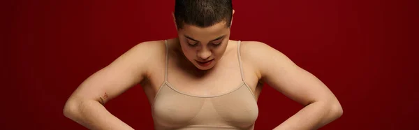 Body positive, self-esteem, young woman with short hair and tattoo posing with hands on hips on burgundy background, dark red, curvy fashion, comfortable in skin, female underwear, banner — Stock Photo