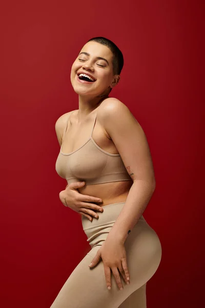 Body positive, happy and tattooed woman in beige underwear posing on red background, curvy fashion, comfortable in skin, body positivity, generation z, body diversity, laughter, joy — Stock Photo
