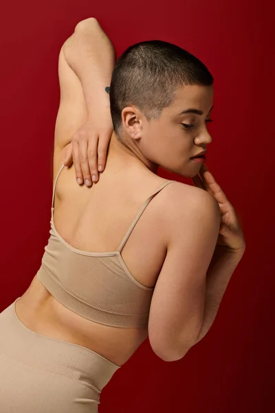 Тіло позитивно, комфортно в шкірі, татуйована молода жінка з коротким волоссям та татуюванням позує з рукою позаду бордового фону, темно-червона, пишна мода, жіноча білизна — Stock Photo